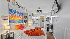 Habitación de hotel moderna con una cama grande, un cuadro sobre la cama, un escritorio con una lámpara y un baño visible a través de una puerta abierta.