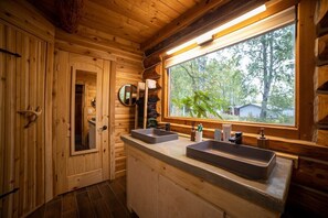 A wooden cabin bathroom with two sinks, a large window, and a mirror. - The Airstrip (Talkeetna)