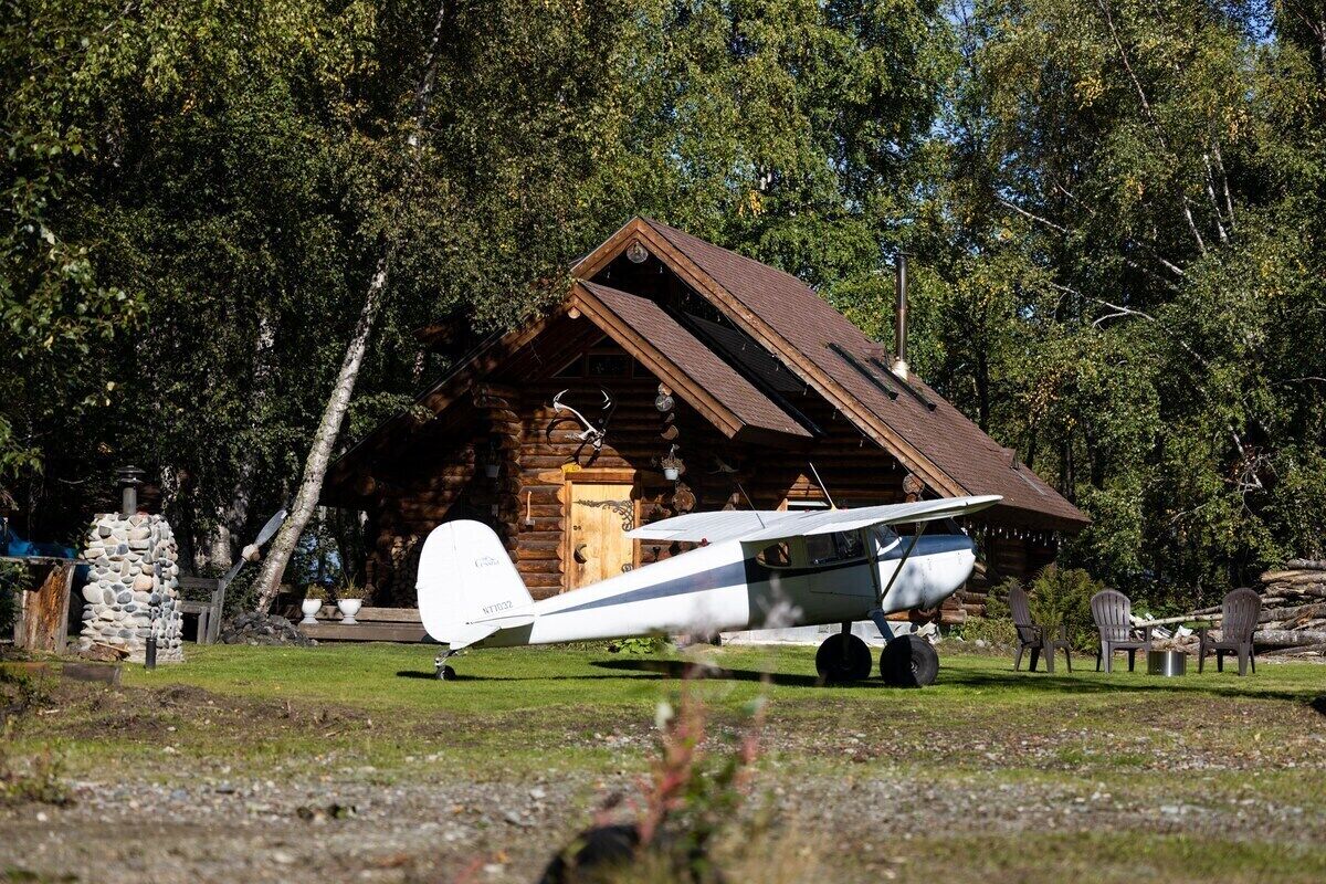 Un pequeño avión blanco estacionado frente a una cabaña rústica de troncos con techo inclinado.