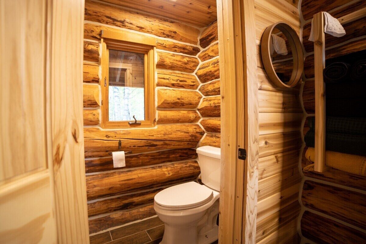 Un baño de cabaña de troncos con interior de madera, un inodoro, una ventana y un espejo.