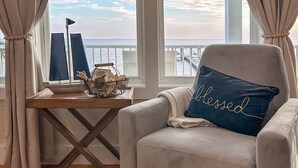A room with a view of the sea, a chair with a pillow, a table with a lamp, and a window with curtains. - “Beach’d Pearl” Beach house with Private Beach and Pier in Ocean Springs (Ocean Springs)