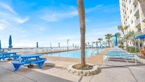 A poolside area with blue picnic tables, a palm tree, and a building with balconies. - NEW! Tide's End Getaway (Panama City Beach)