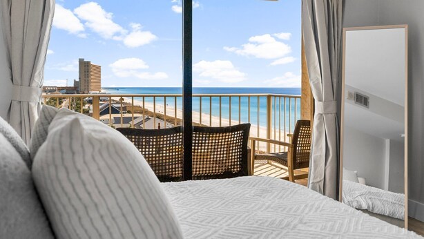 A balcony with a view of the beach and ocean, a bed with white bedding, and a wooden chair. - NEW! Tide's End Getaway (Panama City Beach)