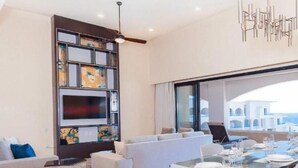 Un salon moderne comprenant un canapé, une table à manger en verre, un ventilateur de plafond et une grande fenêtre donnant sur des immeubles.