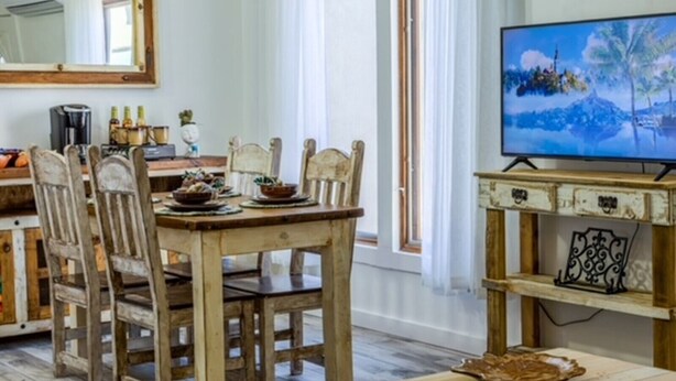 Un área de comedor con una mesa y sillas de madera, un televisor sobre un mueble de madera y una ventana con cortinas.