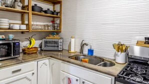 Una cocina con microondas, tostadora, lavaplatos y estufa. Hay estantes con vajilla y un bol de fruta.