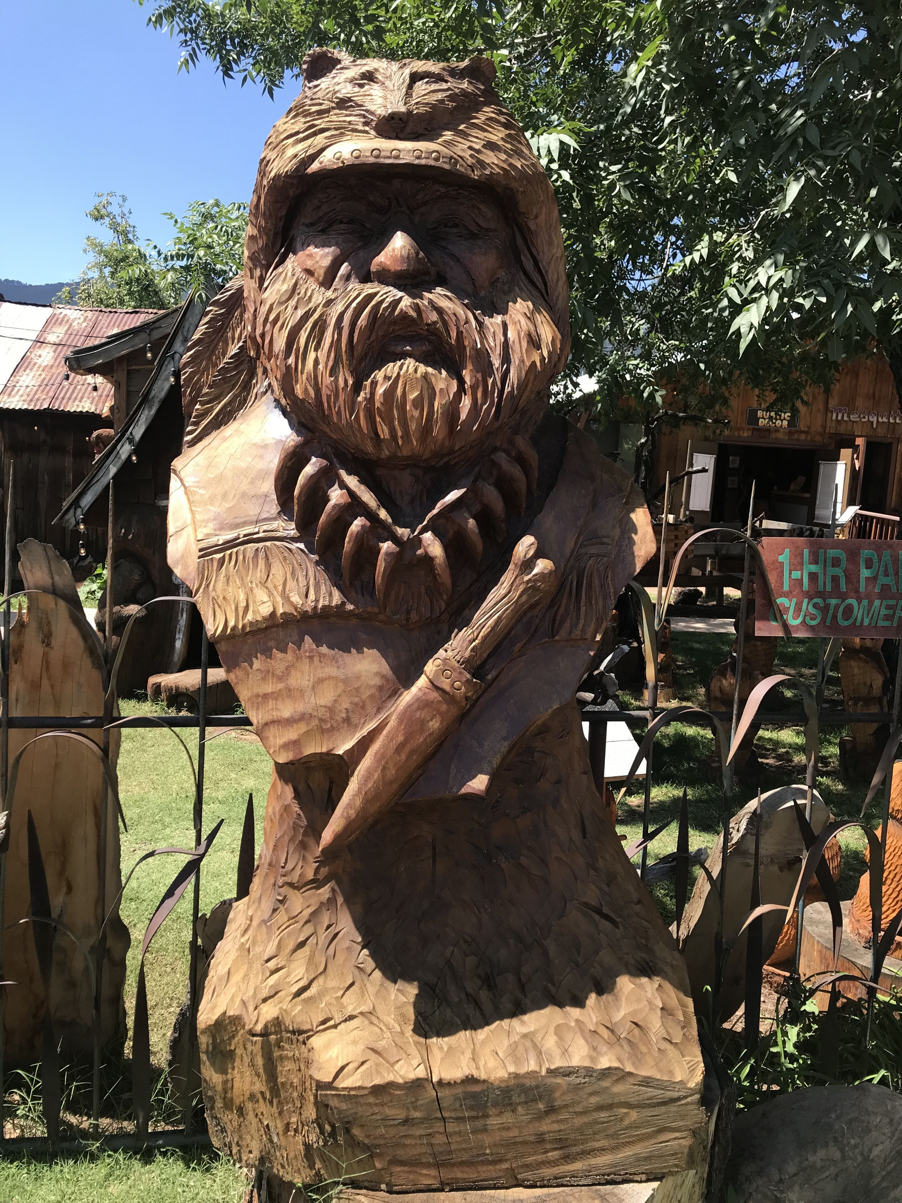 A wooden sculpture of a bearded man holding a stick, surrounded by rustic buildings and greenery.