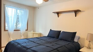 A bedroom with a bed, a nightstand, a ceiling fan, a window with sheer curtains, and a wall-mounted shelf.