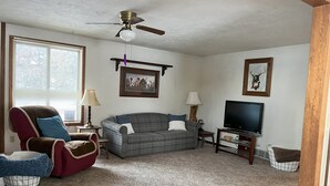 A living room with a red armchair, a grey couch, a ceiling fan, a television, and framed pictures on the walls.