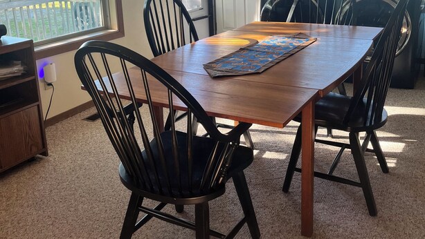 A dining area with a wooden table, four chairs, a hanging light fixture, and a view of a grassy yard through a window.