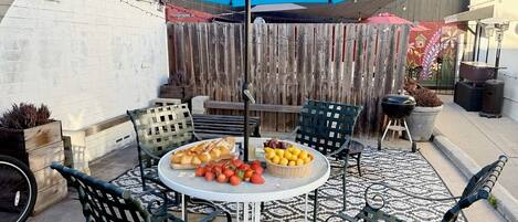 A patio with a round table, four chairs, a blue umbrella, and a wooden fence in the background.