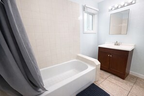 Un baño con bañera, tocador con lavabo, espejo y ventana con persianas.