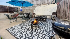 Un patio con una fogata, mobiliario exterior y una sombrilla azul.