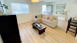 Sala de estar con un sofá color beige, una mesa de centro y un mueble para televisión. Se ve una ventana con persianas, una puerta y una lámpara sobre una mesita auxiliar.