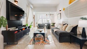 Een moderne woonkamer met een flatscreen televisie, een zwarte bank, een salontafel en een eethoek op de achtergrond.