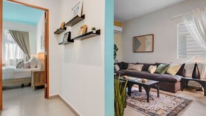 A modern living room with a sofa, coffee table, and decorative items. There is a bed with a nightstand and a lamp. The room has a light blue accent wall and a large window with curtains. - Stylish Apartment in Condado (San Juan)