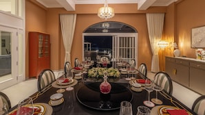 Een eetkamer met een ronde tafel gedekt voor een maaltijd, een kroonluchter en een uitzicht op buiten door een groot raam.
