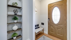 Un pasillo con puerta de madera, un banco de bienvenida y estantes con plantas y adornos.