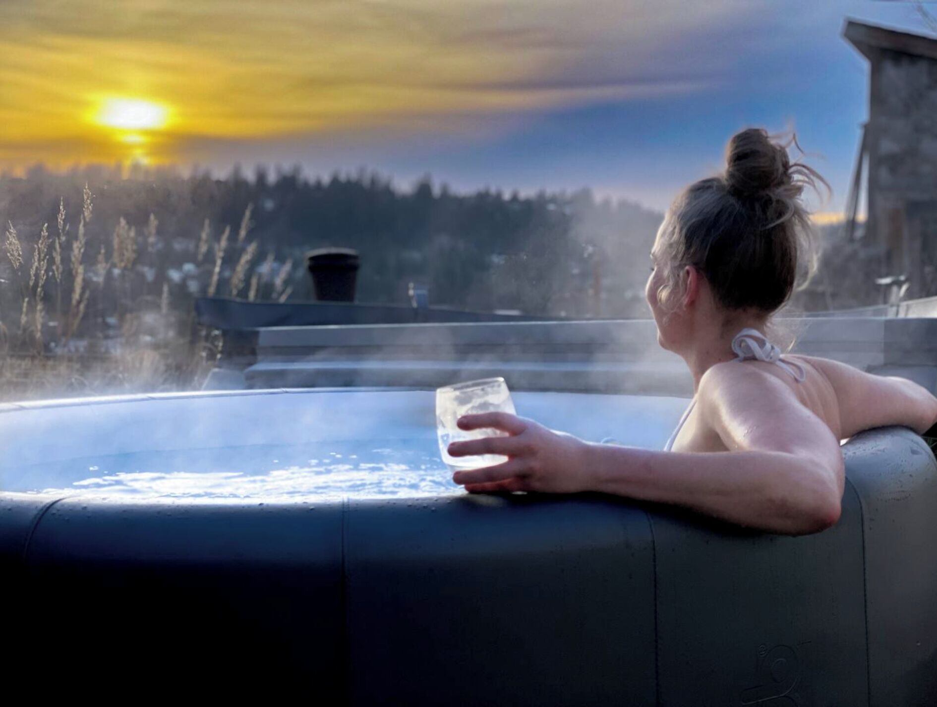 Une personne se détend au bord d’un jacuzzi, un verre d’eau à la main, et contemple une zone boisée au coucher du soleil.