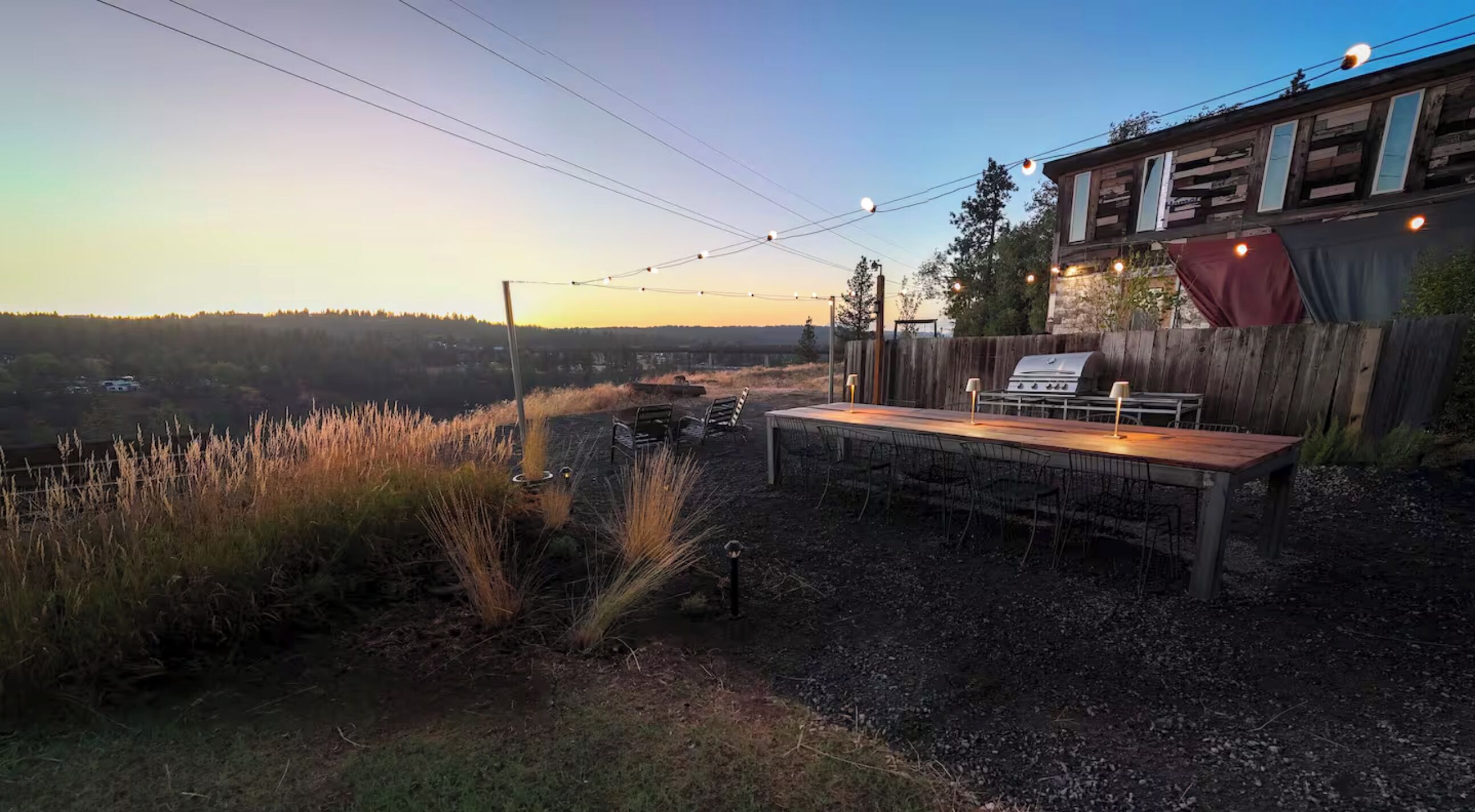Un espacio de comedor al aire libre con una mesa y sillas de madera, luces colgantes y una parrilla.