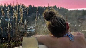 A person relaxing in a hot tub with a glass of drink, overlooking a forested area. - Sunset Ridge, Near Downtown, Hot tub, Outdoor Living, all with Panoramic View,  (Spokane)