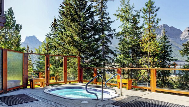 Een hottub met een houten terras en metalen reling, omringd door bomen en bergen op de achtergrond.