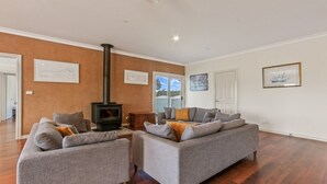 A spacious living room with a large grey sofa set, a black wood-burning stove, and a sliding glass door leading to a balcony. - Peninsula at Metung House | Boat Access Only Getaway (Boole Poole)