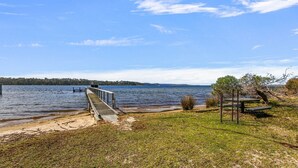 A lakeside view with a wooden dock extending into the water, surrounded by grassy areas and trees. - Peninsula at Metung House | Boat Access Only Getaway (Boole Poole)