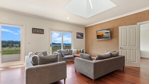 A spacious living room with a large window offering a view of the outdoors, a sofa set, and a painting on the wall. - Peninsula at Metung House | Boat Access Only Getaway (Boole Poole)