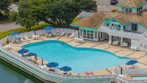Ein Schwimmbecken mit Liegestühlen und Sonnenschirmen, ein Gebäude mit Ziegeldach und eine eingezäunte Terrassenfläche.