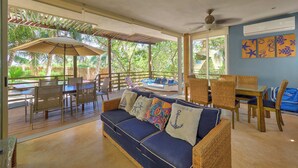 A spacious room with a large sofa, dining table, and chairs. There is a balcony with a view of palm trees and a wooden deck.
