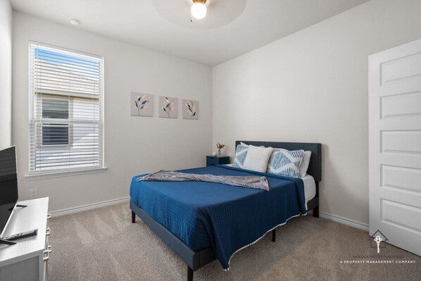 A bedroom with a bed, a desk, a window with blinds, and a door.