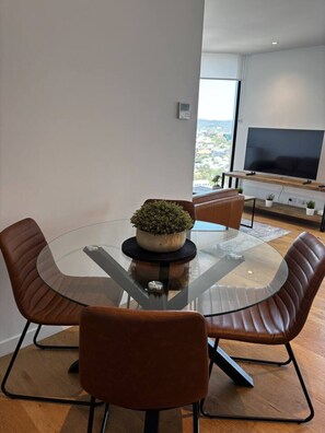A modern dining area with a glass table, leather chairs, a potted plant, and a TV in the background. - Luxury Peppers Infinity Pool +Stylish 2BD+1C Park (Fortitude Valley)