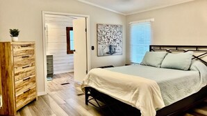 Um quarto com cama, ventilador de teto, cômoda de madeira, quadro e janela com persianas.