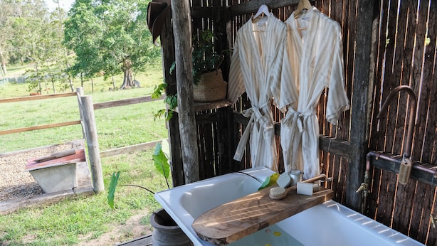 A rustic outdoor bathroom with a wooden tub, a wooden bench, and a hanging clothes rack.