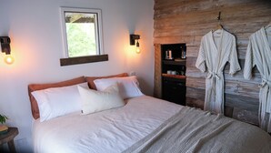A bed with white linens, two wall-mounted light fixtures, a window, and a wooden wall with a robe hanging.