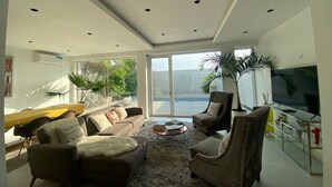 A modern living room with a sofa, armchairs, a coffee table, and a TV. There is a large window with a view of a body of water and greenery outside. - Modern Beachfront Luxury in Monrovia  (Monrovia)