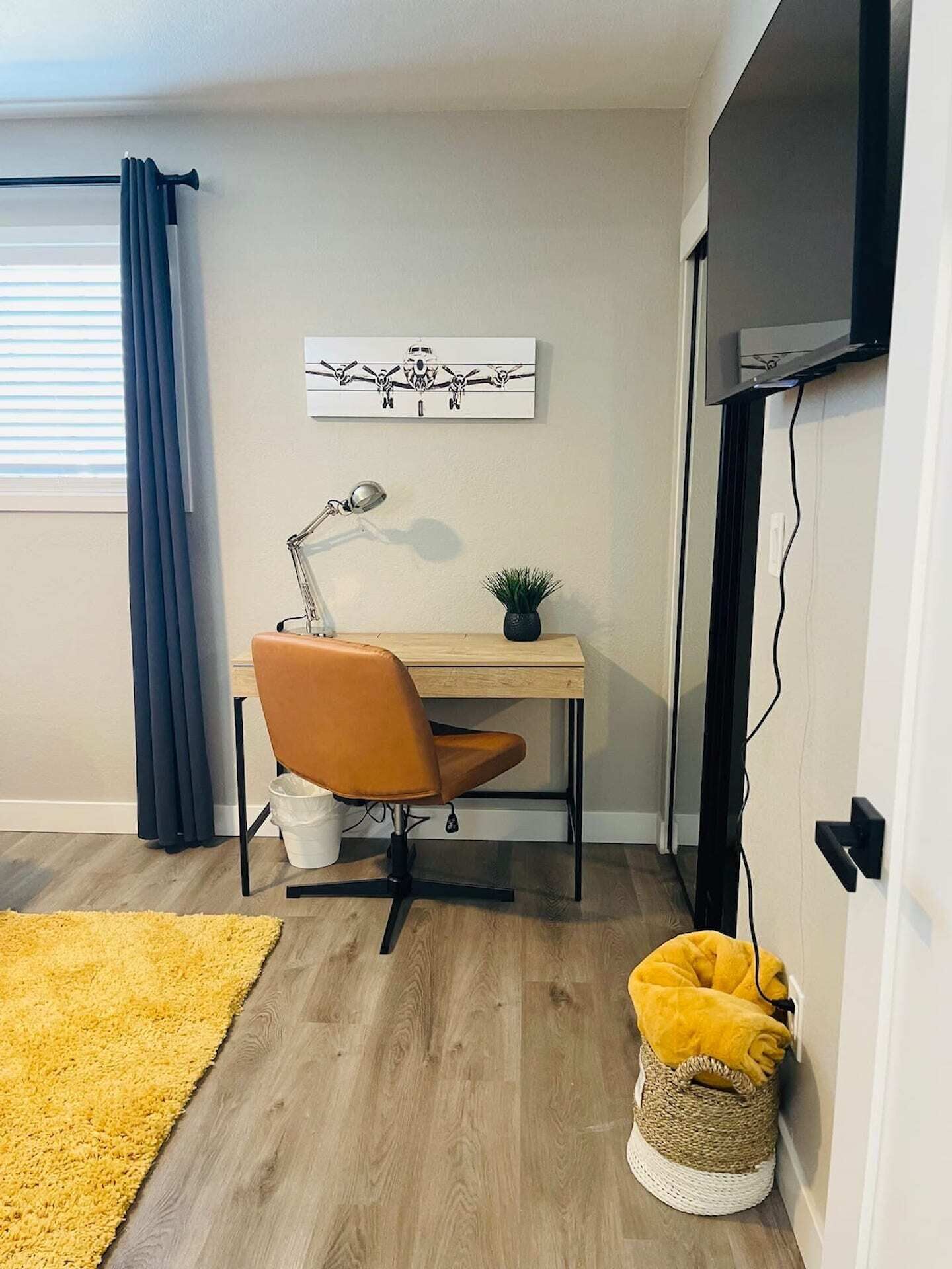 Un espacio de oficina moderno con un escritorio de madera, una silla naranja, una lámpara de escritorio, una pequeña planta en maceta, una alfombra amarilla y una cesta tejida.
