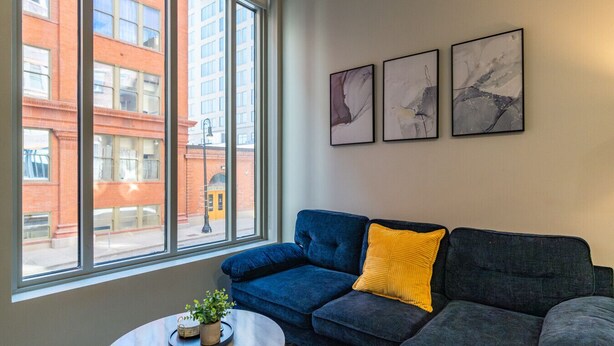 A modern living room with a blue sofa, a yellow pillow, and a small table with a plant. Three abstract paintings are on the wall. - 2Bedroom, Near Over theRhine & Fountain Sq. + wifi (Cincinnati)