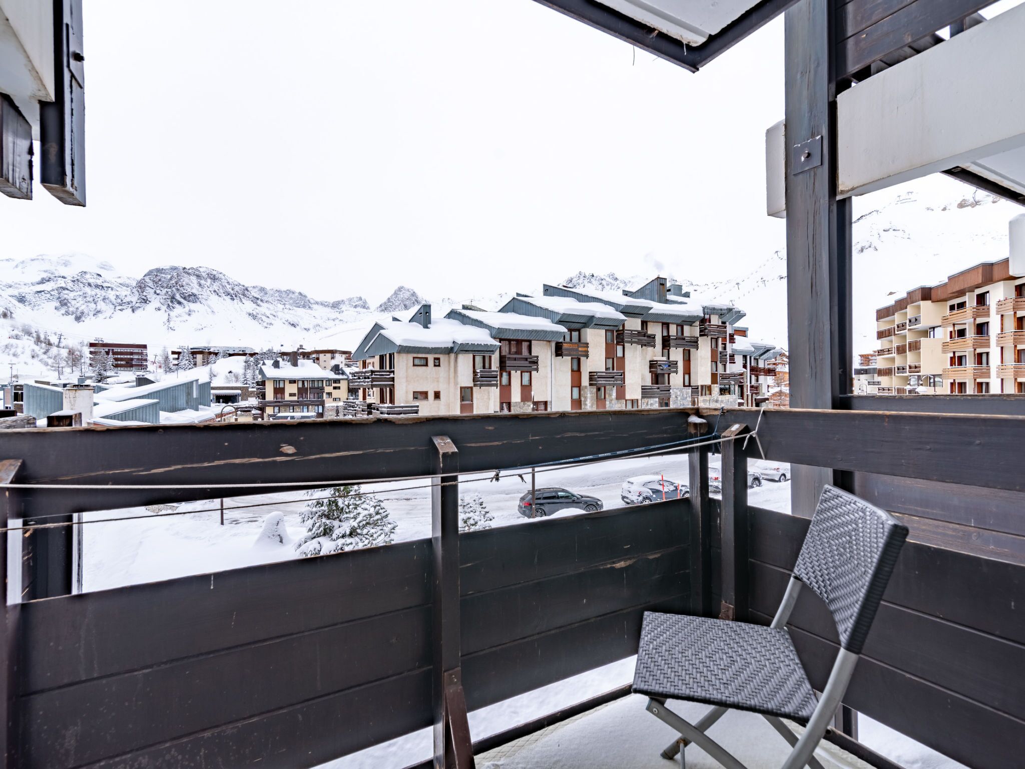 A balcony with a chair, overlooking a snowy mountain town with buildings and a clear sky.
