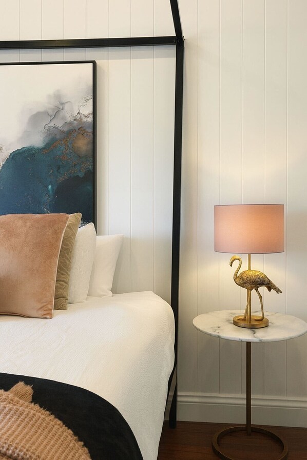 Una cama bien hecha con cubrecama blanco y varios almohadones, un cuadro abstracto enmarcado sobre la cama, y una pequeña mesa redonda con una lámpara que tiene un diseño de flamenco dorado.