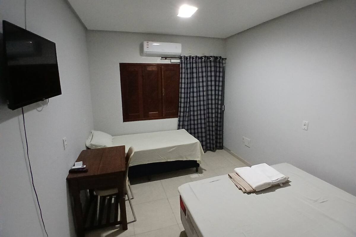 A small, clean room with two beds, a desk, a chair, a TV, and an air conditioning unit.