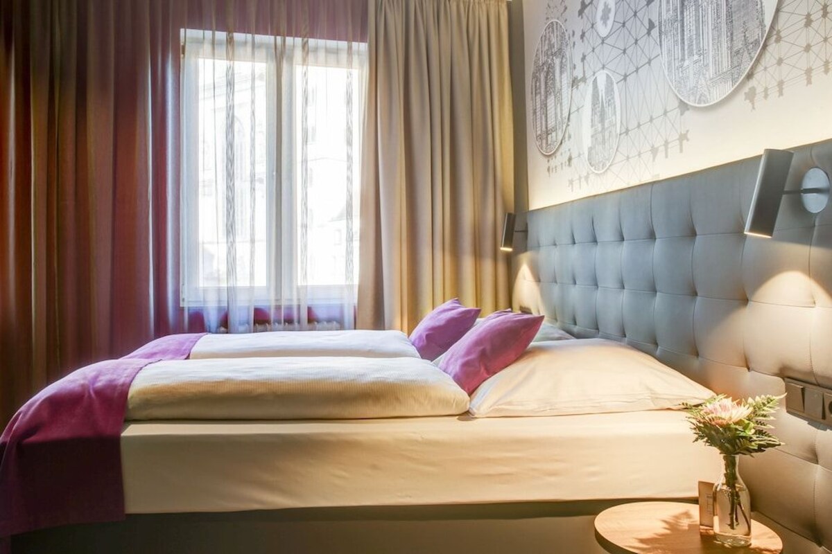 A double bed with a headboard, a bedside table with a vase, and a window with curtains.