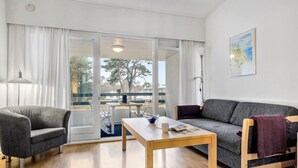 Un salon moderne avec un canapé gris, une table basse en bois et un fauteuil gris. Une grande fenêtre est agrémentée de rideaux blancs, un tableau orne le mur et offre une vue sur l’extérieur.