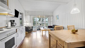 Une cuisine moderne avec des appareils électroménagers blancs, un coin repas avec une table et des chaises en bois, et une vue sur l’extérieur grâce à de grandes fenêtres.