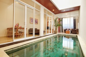 A modern indoor pool with glass walls, a wooden ceiling, and a dining area with chairs and a table.
