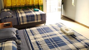 Una habitación con dos camas, cada una con colcha a cuadros azules y blancos, una cortina verde y amarilla, y una mesita con un control remoto.
