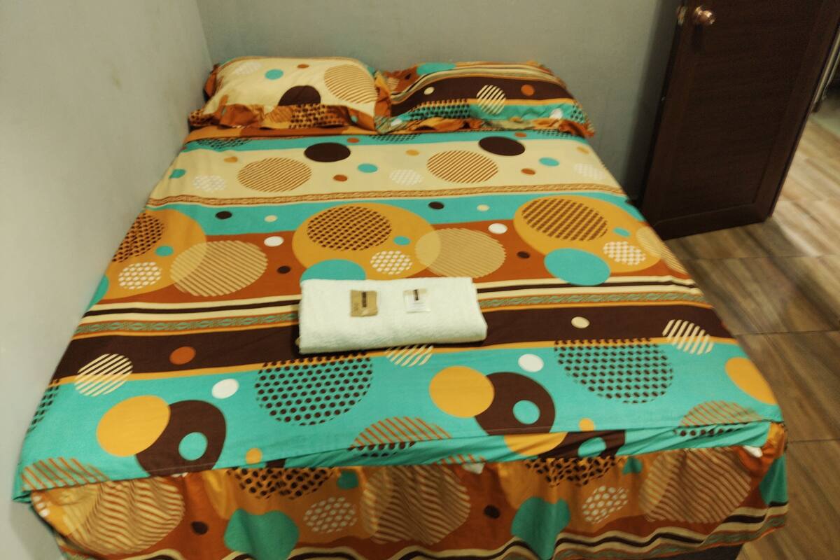 Una cama con una colcha estampada y almohadas, una manta blanca y una pequeña bolsa blanca sobre la cama.