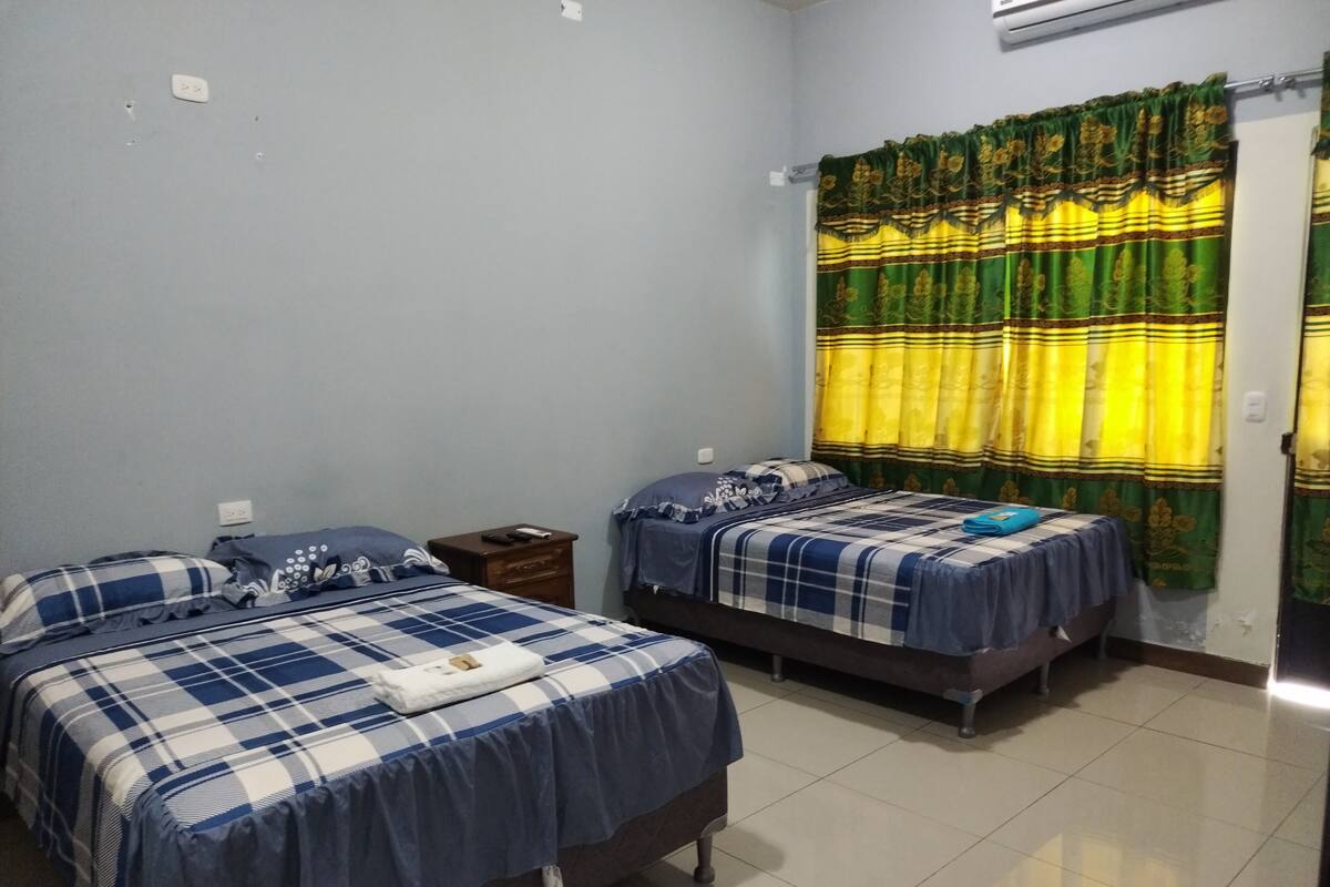 Una habitación con dos camas, cada una con una colcha a cuadros azules y blancos, una mesita de noche y una ventana con cortinas verdes y amarillas.