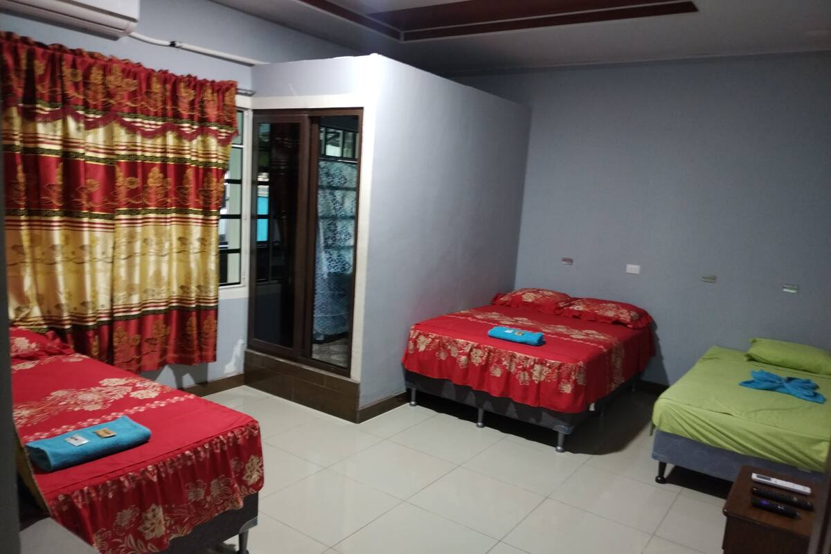 Habitación con dos camas, una con cubrecolcha rojo y la otra con cubrecolcha verde. Se ve una ventana con cortinas rojas, una puerta y una mesita con un control remoto.